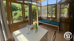 A sunlit room with a table, a bench, and large windows offering a view of greenery.