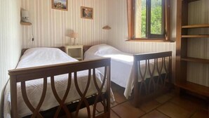 A room with two beds, wooden beams on the ceiling, a window with a view of greenery, and a small shelf with a lamp.