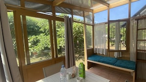 A sunlit room with a table, a bench, and large windows offering a view of greenery.