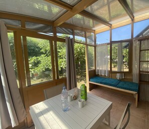 A sunlit room with a table, a bench, and large windows offering a view of greenery.