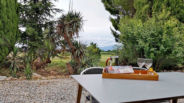 Mesa exterior con una bandeja que contiene una jarrón, dos copas de vino y un menú, sobre un fondo de grava, árboles y una vista distante de montañas.