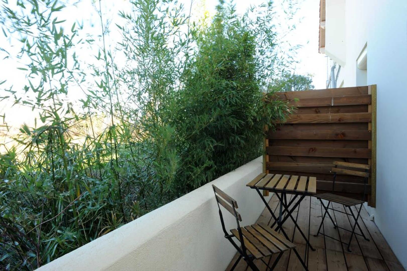 A small outdoor seating area with a wooden table and chairs, surrounded by dense foliage and a white wall.