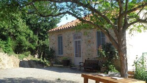 Un bâtiment rustique en pierre, avec un toit en tuiles, un banc et un arbre au premier plan.