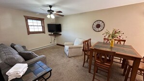 Un salon comprenant un canapé, une table basse, une table à manger avec des chaises, un ventilateur de plafond, un téléviseur et une horloge murale.