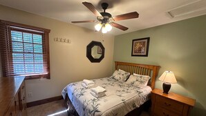 Une chambre à coucher comprenant un lit, une table de chevet, une lampe, un ventilateur de plafond, une fenêtre avec des stores, un miroir et un tableau accroché au mur.