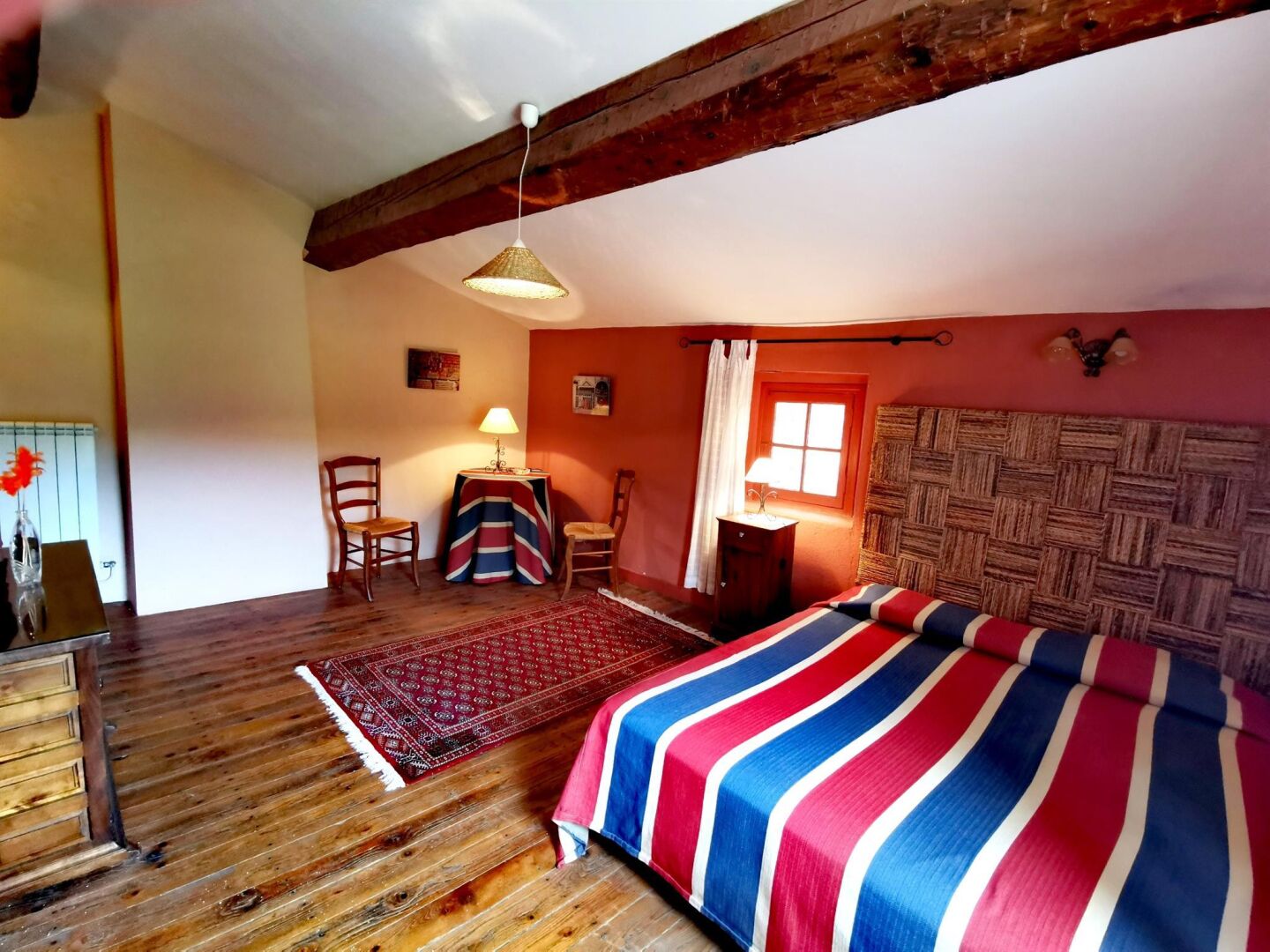 A bedroom with a wooden floor, a bed with a striped blanket, a wooden headboard, a small table with a lamp, and a window with curtains.