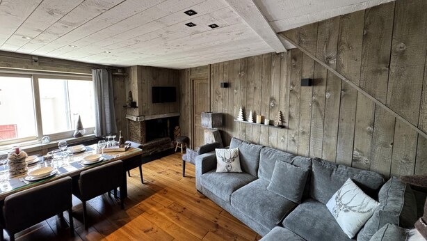 A cozy living room with a fireplace, a grey sofa, and a dining area with a table set for four. - Gîte Sylvestre - LLO - Gîte (LLO)