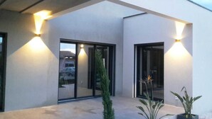 Un edificio moderno con amplias puertas y ventanas de vidrio, iluminación exterior y plantas en macetas.