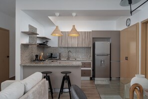 A modern kitchen with granite countertops, a built-in oven, and a stainless steel refrigerator. There are two bar stools and a bench in the dining area.