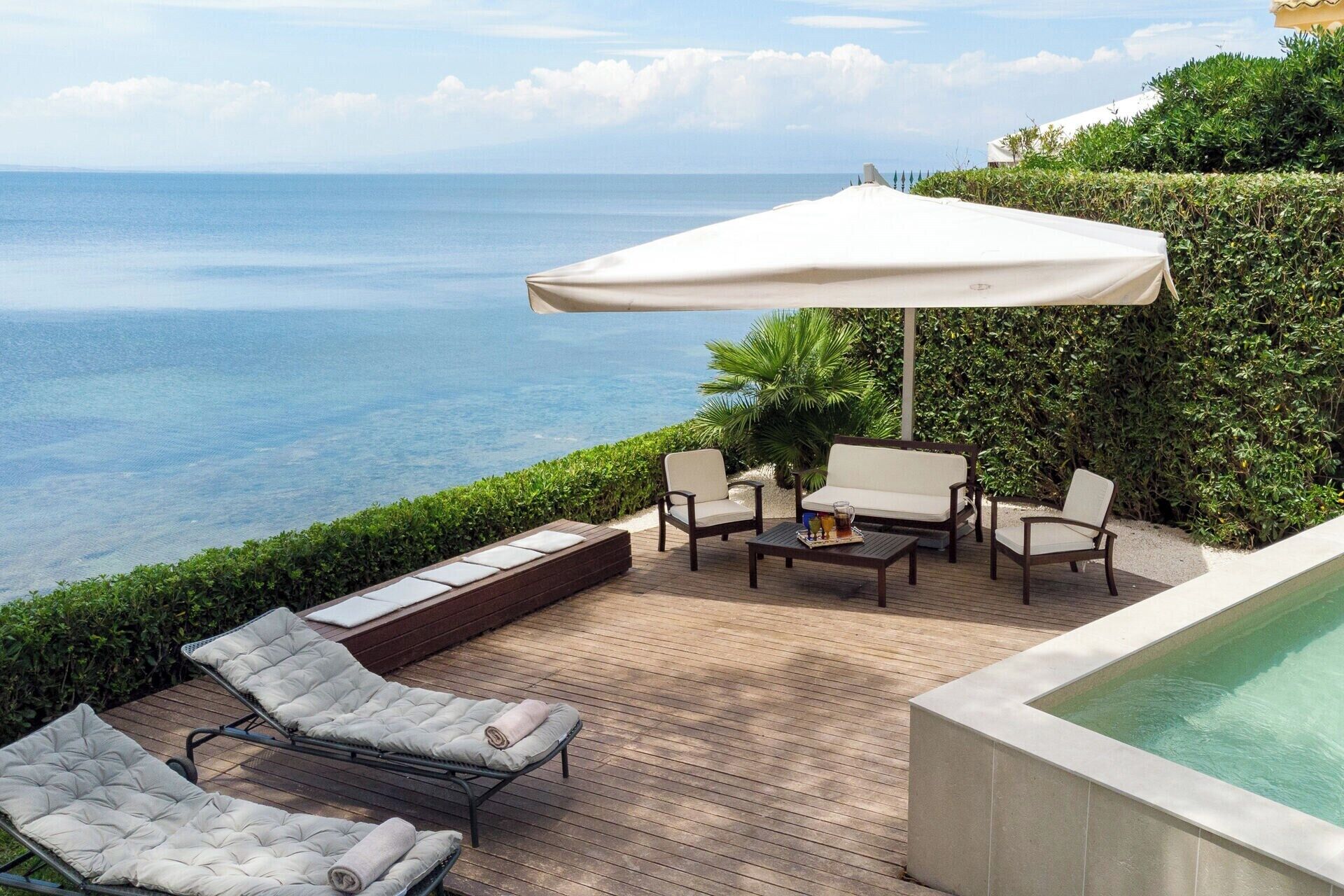 A deck with lounge chairs, a table, and an umbrella overlooking a pool and the sea.