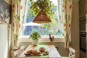 Eine Küche mit Holztisch, Stühlen, einer Hängeleuchte, einem Fenster mit Vorhängen und Blick auf Grünfläche.