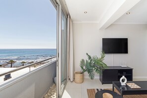 A modern living room with a large window overlooking a beach, a flat-screen TV mounted on the wall, and a black entertainment unit.