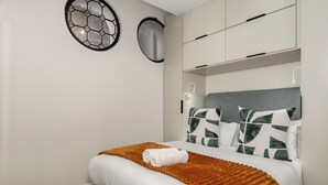A neatly made bed with a white comforter and a brown throw blanket, a headboard with patterned pillows, and a wall-mounted light fixture.