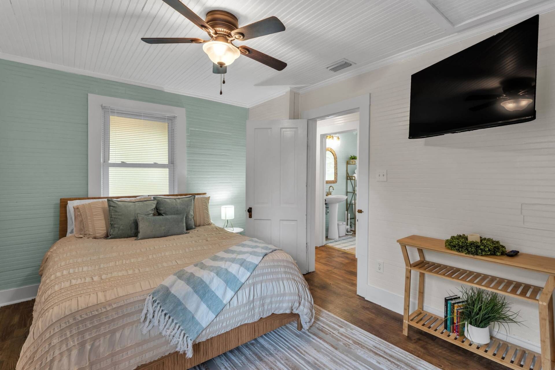 A bedroom with a bed, a ceiling fan, a TV mounted on the wall, and a console table with plants.