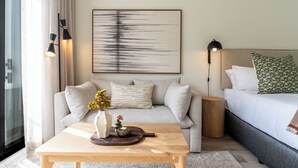 A modern hotel room with a sofa, a wooden coffee table with a vase and decorative items, a bed with pillows, and a large abstract painting on the wall. - The York | Designer CBD Apartment (Cape Town)