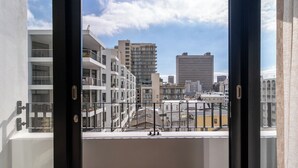 A view from a window with a metal railing, overlooking a cityscape with various buildings, including a tall, modern structure and a shorter, multi-story building with balconies. - The York | Designer CBD Apartment (Cape Town)