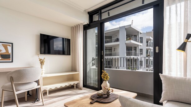 A modern living room with a sofa, a wooden coffee table, a TV mounted on the wall, and a balcony with a view of the building's exterior. - The York | Designer CBD Apartment (Cape Town)