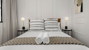 Una cama bien hecha con dos almohadas, dos lámparas de noche y dos toallas enrolladas sobre un edredón estampado.
