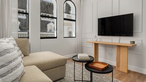 Un salón moderno con un sofá, una mesa de centro, un televisor montado en la pared y una ventana con vistas a un edificio.