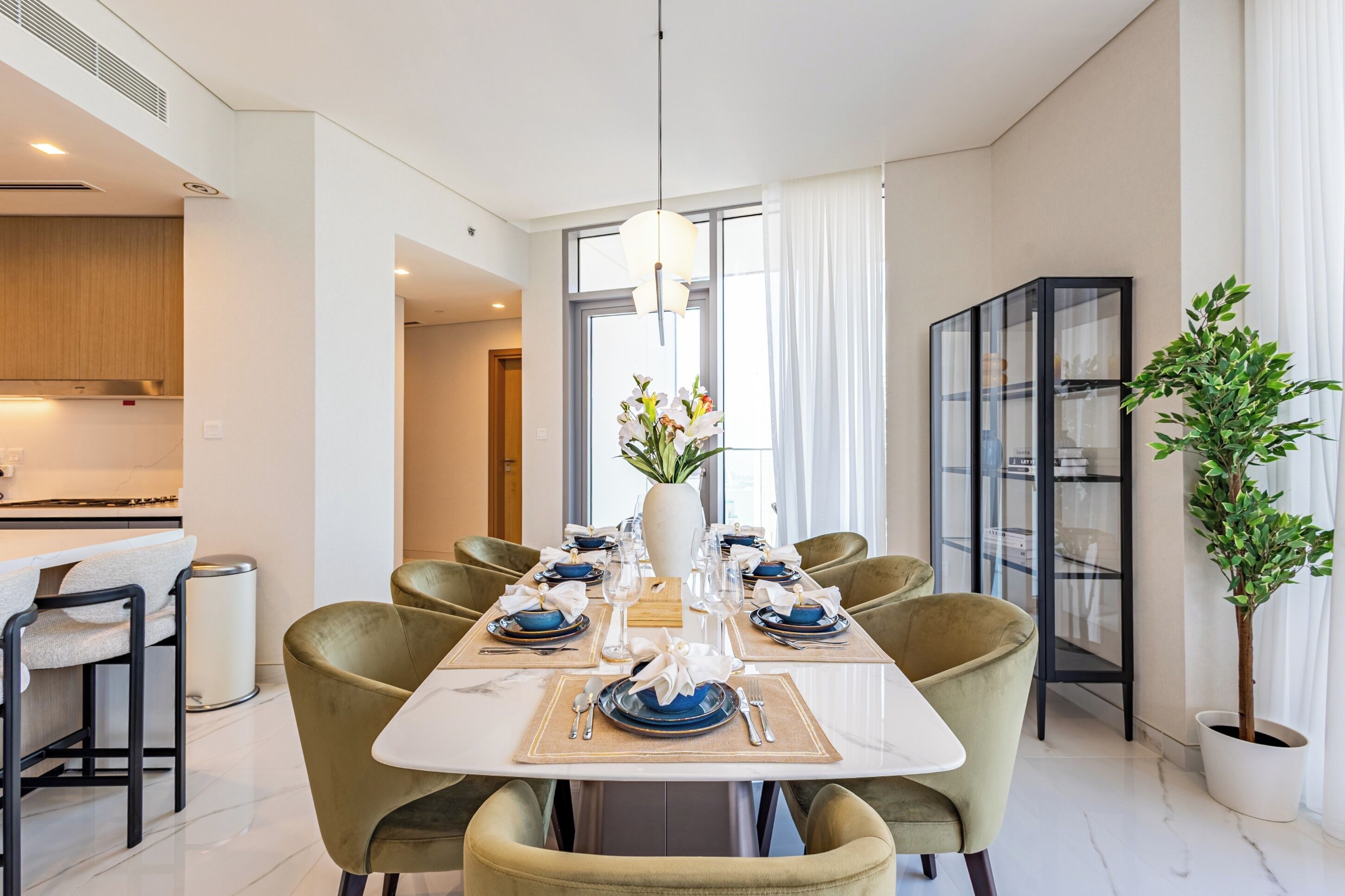 Un comedor moderno con una mesa blanca dispuesta para seis personas, rodeada de sillas tapizadas de color verde. Un jarrón con flores está centrado en la mesa, y se ve un aparador de vidrio a la derecha.