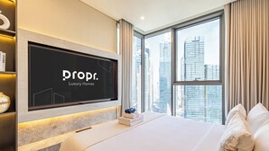 Ein modernes Hotelzimmer mit einem großen Fernsehbildschirm, der 'prop. Luxury Homes' zeigt, einem ordentlich bezogenen Bett, Blick auf die Stadt und einem Bücherregal mit dekorativen Gegenständen.