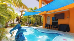 Una piscina con una escultura de delfín, asientos al aire libre y una vela de sombra azul.