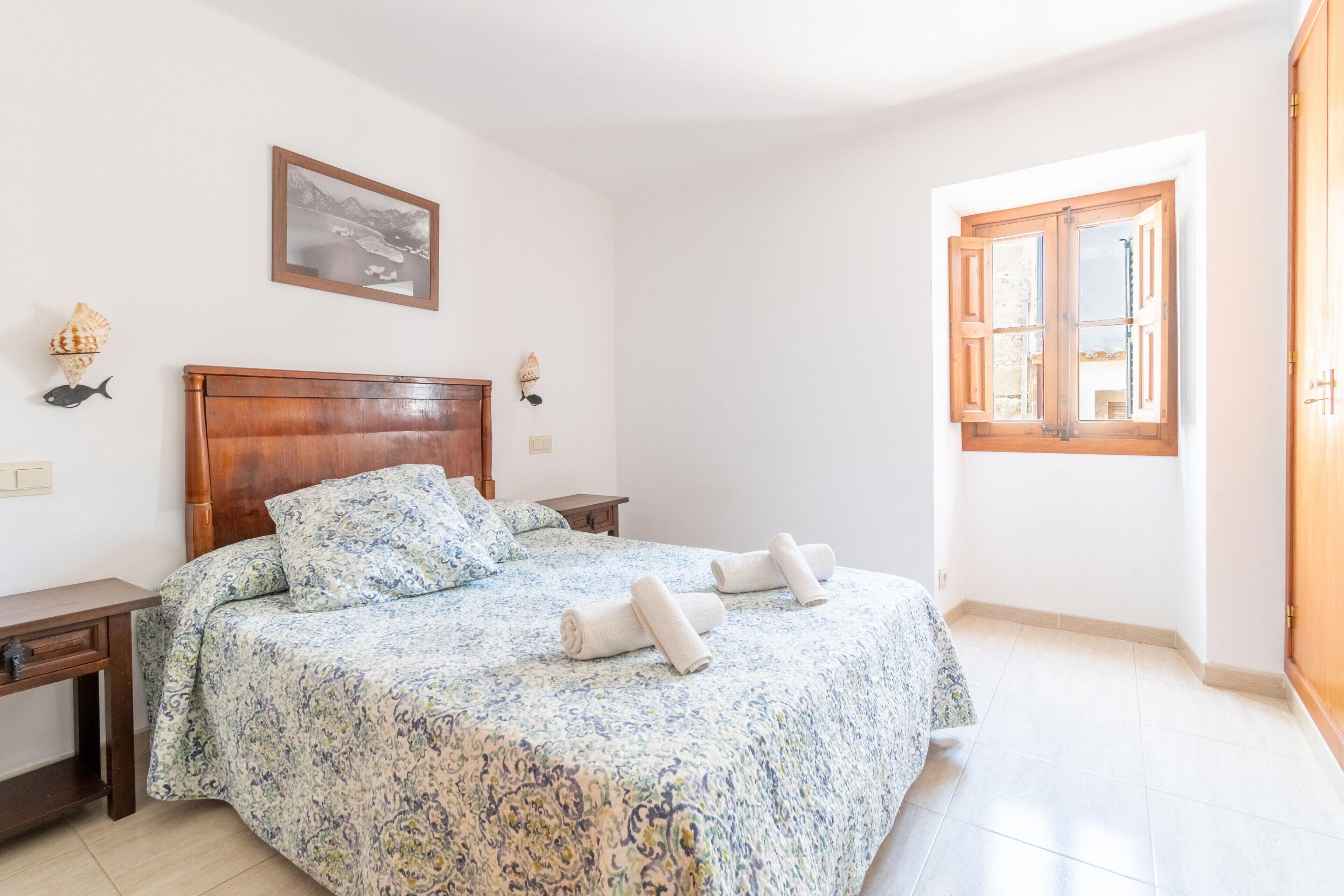 Een slaapkamer met een houten bed, een raam met houten luiken, een nachtkastje en een aan de muur bevestigd schilderij.