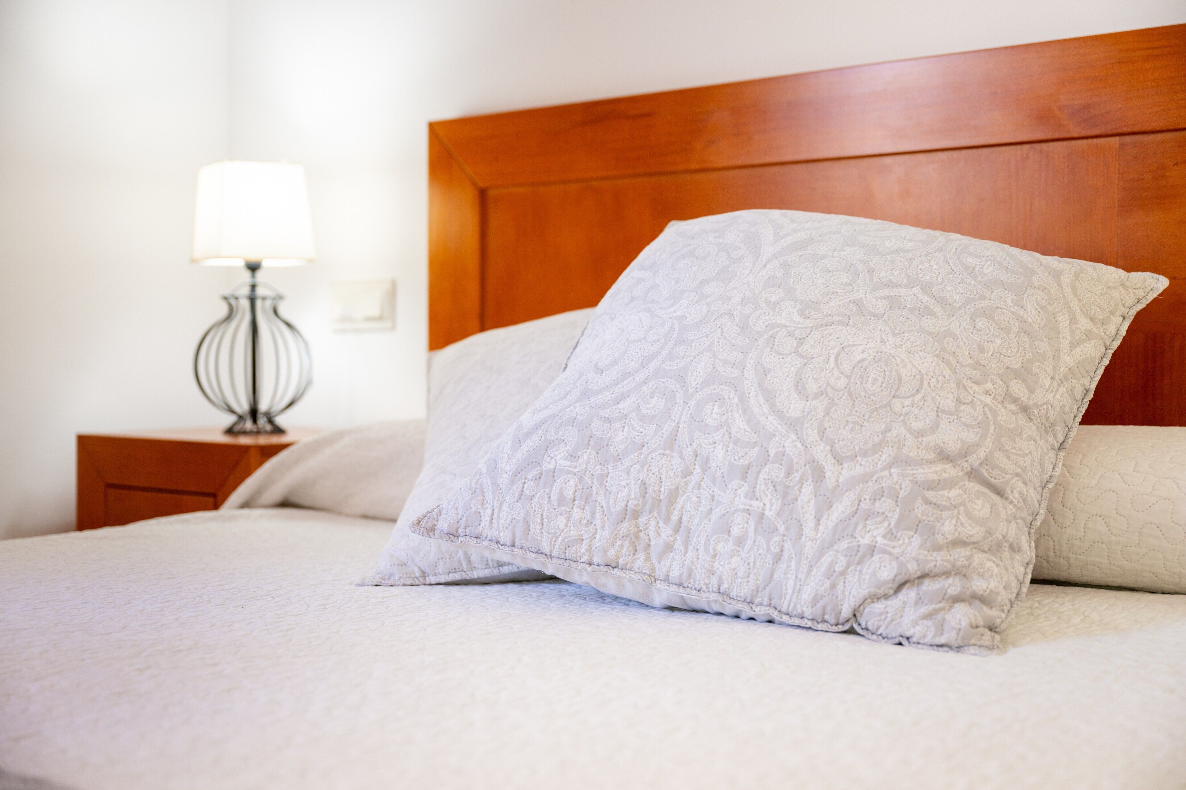 A neatly made bed with a wooden headboard, a bedside lamp, and a patterned pillow.