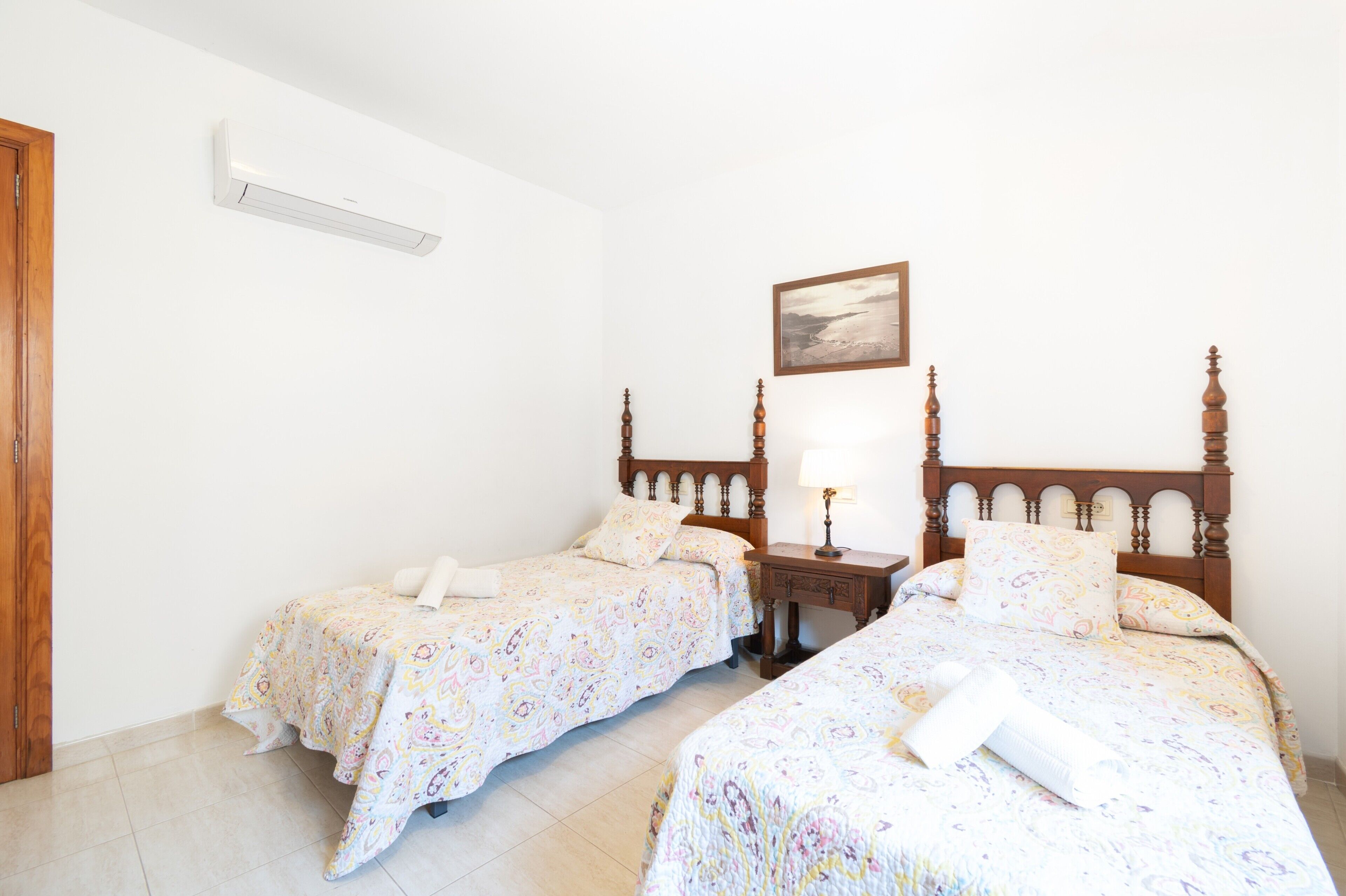 Habitación de hotel con dos camas individuales, cabecero de madera, mesita de noche con lámpara y un cuadro en la pared.