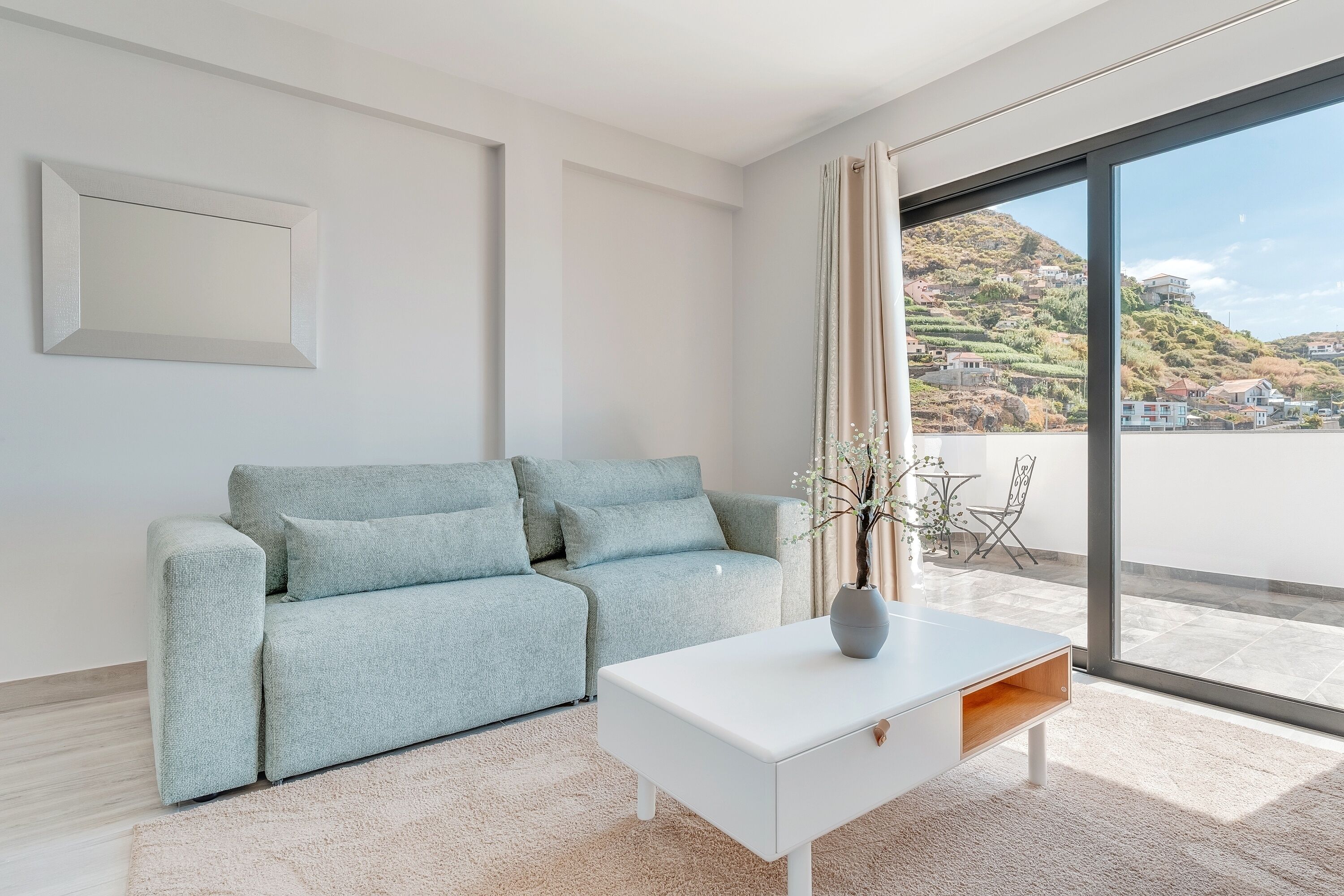 A modern living room with a light blue sofa, a white coffee table, and a large window offering a view of a hillside.