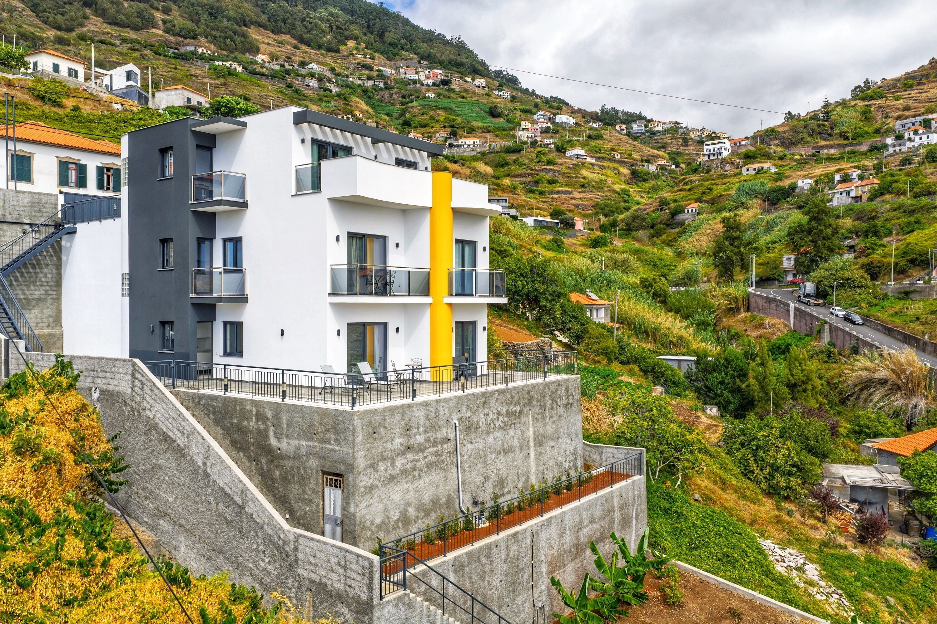 A modern multi-story building with balconies, situated on a hillside with surrounding houses.