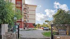 Une entrée fermée par un portail mène à un immeuble résidentiel à la façade rouge et beige, doté d’un balcon visible et d’une allée pavée menant à un parking.