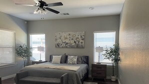 Quarto com cama, mesas de cabeceira, ventilador de teto e duas janelas com persianas.