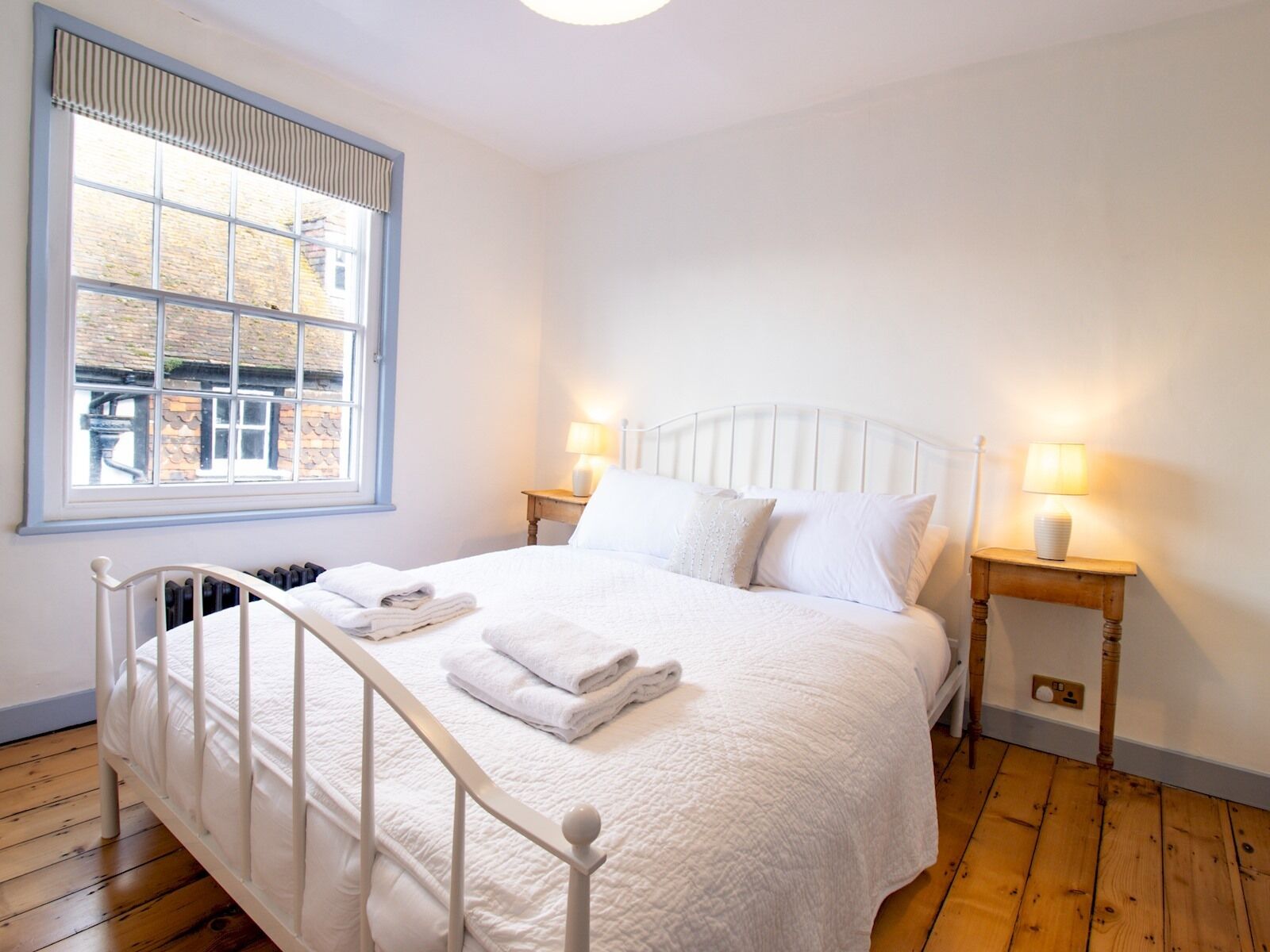 Un letto bianco con poggiatesta, due comodini con lampade e una finestra con vista sugli edifici.