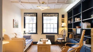 A cozy living room with a sofa, a coffee table, and wicker chairs. There are two windows with black frames and a radiator below them. A bookshelf is visible on the right side. - Easter Cottage - Central Rye, Effortless Chic! | Pass the Keys (Rye)