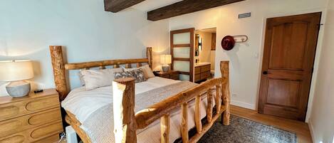 Uma cama de madeira com cabeceira, uma mesa de cabeceira com abajur, cômoda, espelho e uma porta.