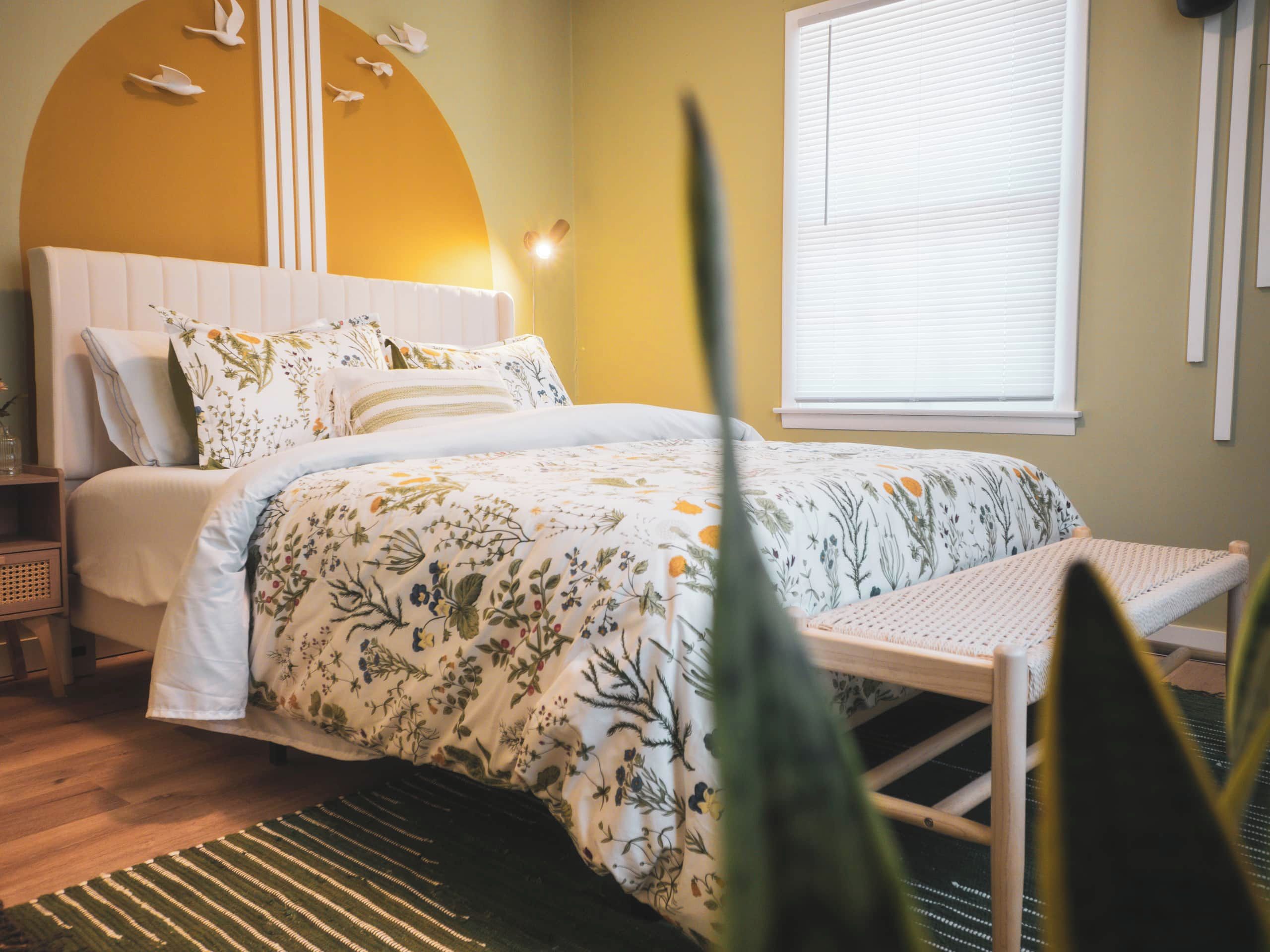 A bedroom with a bed, bedside tables, a window with blinds, and a wall with a curved design and hanging decorations.