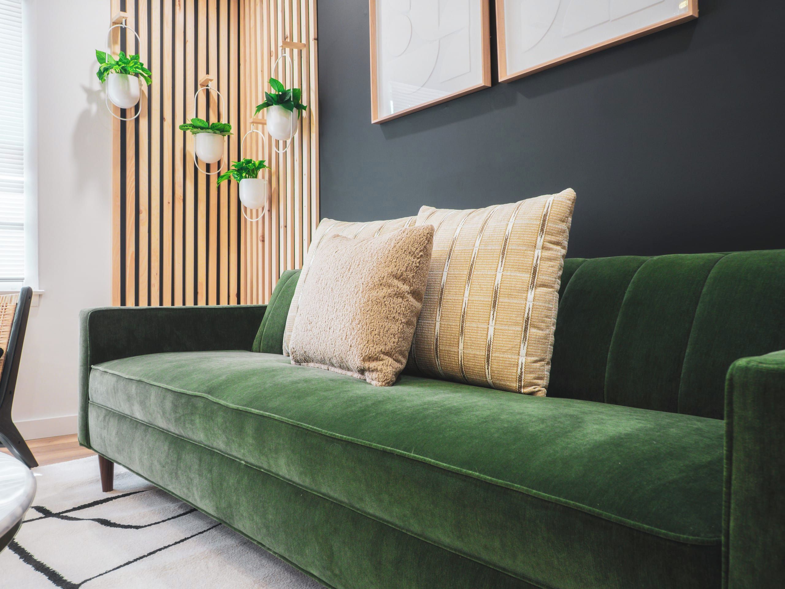 Un sofá de terciopelo verde con dos almohadas, una pared de listones de madera y macetas colgantes.