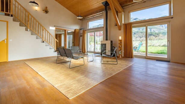 A spacious living area with a wooden floor, a staircase, and large windows offering a view of the outdoors.