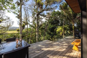 A wooden deck with a glass table and chairs, overlooking a lush landscape with trees and a distant view of hills. - Te Toki Luxury Treehouse and Studio (Waiheke Island)