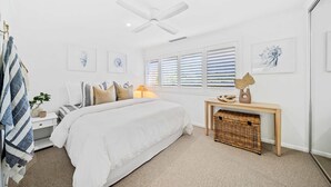 Ein Schlafzimmer mit einem großen Bett, einem Nachttisch, einer Deckenventilator, einem Fenster mit Jalousien und einem kleinen Holzkonsolentisch mit Dekoration.