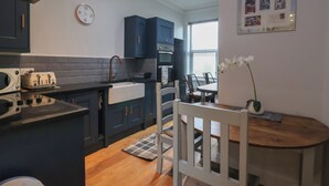 Cocina compacta con armarios oscuros, microondas, tostadora y pileta. Hay un comedor con una mesa y sillas, y un pequeño jarrón decorativo sobre la mesa.
