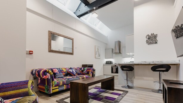 Un saló modern amb un sofà colorit, una taula de centre de fusta i una zona de cuina amb un tamboret.