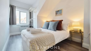 A bedroom with a bed, a bedside table with a lamp, a window with curtains, and a painting on the wall.
