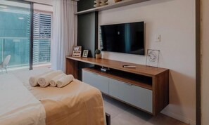 Une chambre d’hôtel moderne, équipée d’un lit, d’un téléviseur posé sur un meuble en bois, d’une petite étagère avec des objets décoratifs et offrant une vue sur l’extérieur.