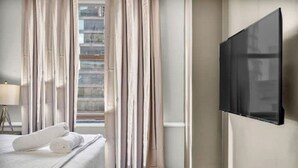 A hotel room with a bed, a TV mounted on the wall, a window with curtains, and a lamp on a side table. - Sophisticated Downtown Loft / FREE Parking & Gym (Cleveland)