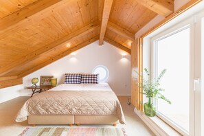 Een slaapkamer met een houten plafond, een bed met een gewatteerde spreideken, een nachtkastje met een lamp en een raam met een plant.