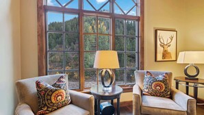 Ein Zimmer mit zwei Sesseln, einem Tisch mit einer Lampe und einem großen Fenster mit Blick auf einen Wald.