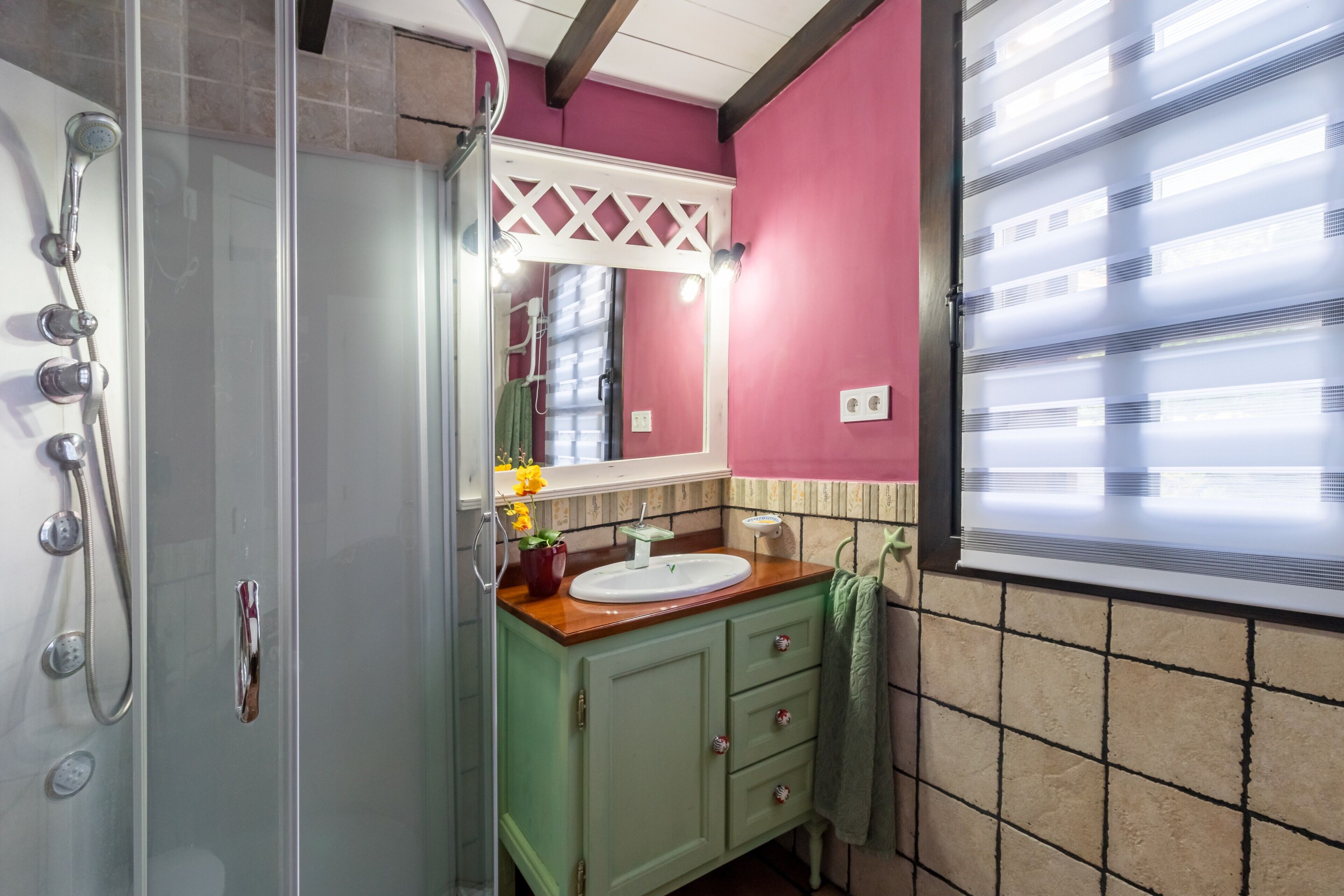 Ein Badezimmer mit Duschkabine aus Glas, einem Waschtisch, einem Spiegel und einem Fenster mit Jalousien.