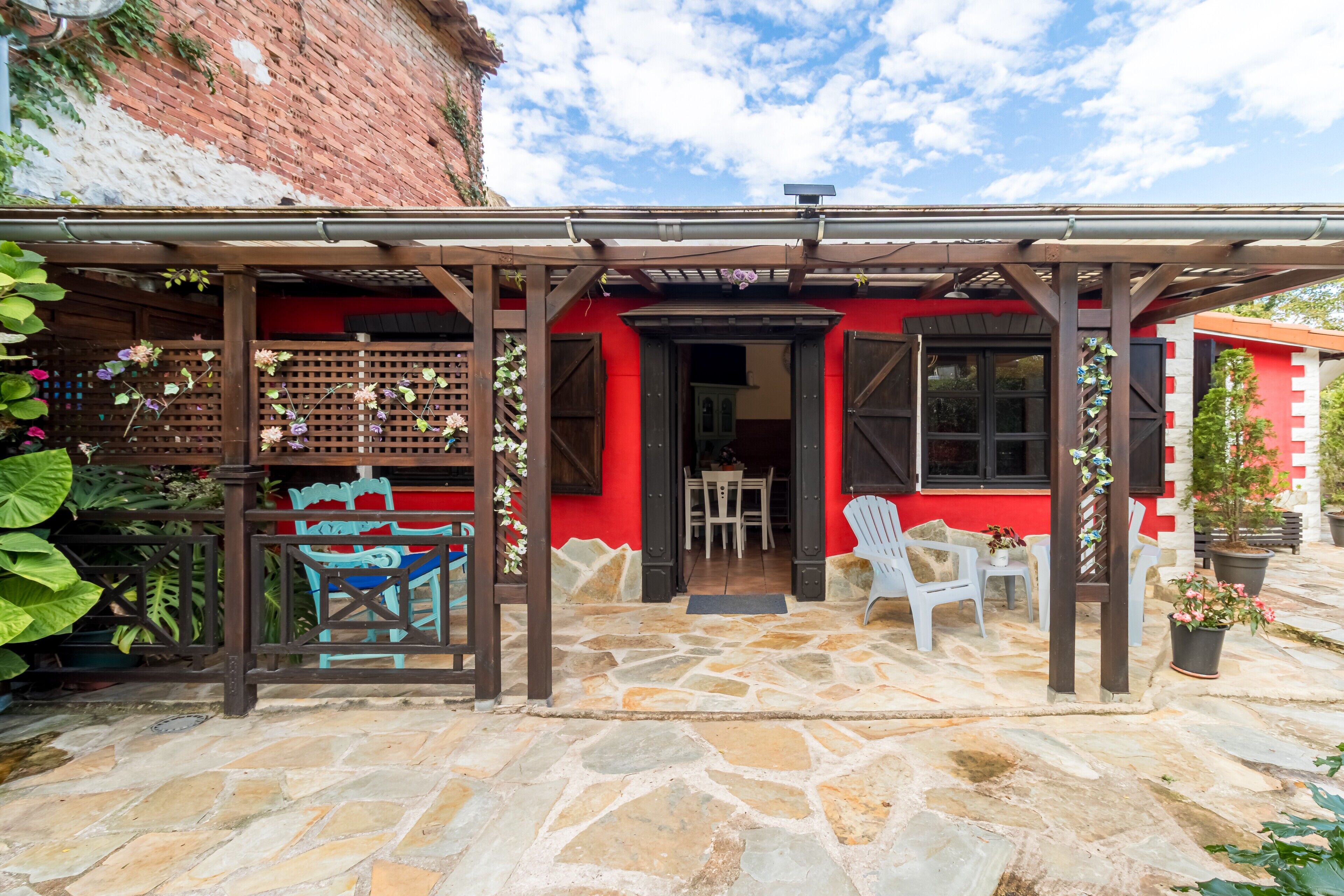 Un patio empedrado con un pérgola de madera, un edificio rojo con una puerta negra y sillas de plástico blanco.